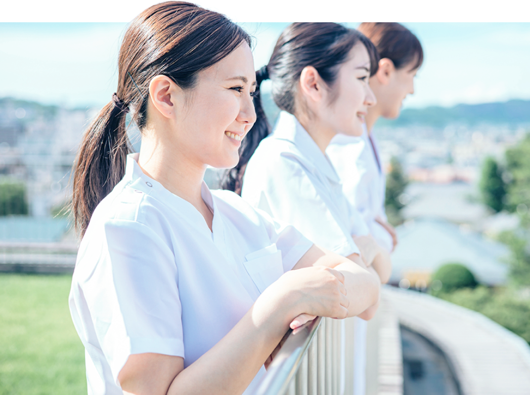 屋外の手すりにもたれ、景色を眺めながら笑顔を交わす白衣の看護師たち