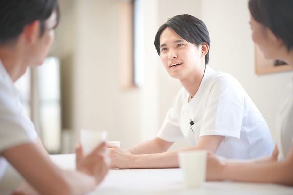 休憩室で紙コップを手に同僚と談笑する白衣姿の若手医療スタッフ